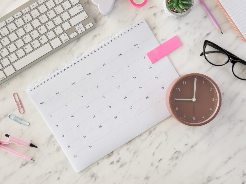 Mesa com calendário e relógio analógico representando a jornada de trabalho e a necessidade de ter um controle de ponto eficiente