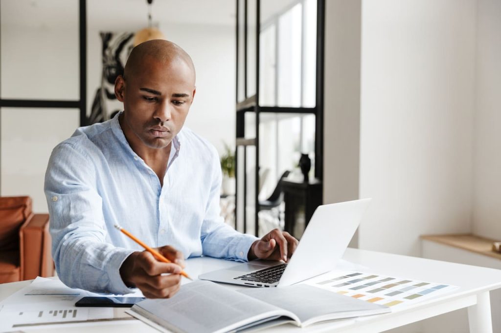 Controle de absenteísmo Foto de um homem afro-americano concentrado escrevendo notas enquanto trabalhava com o laptop na mesa na sala de estar